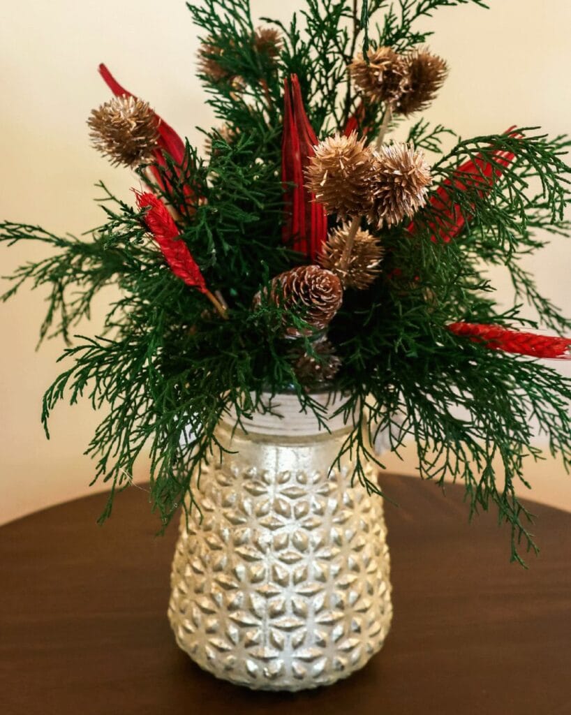 Composition de feuillage vert avec pommes de pin et touches de rouge dans un vase argenté texturé.